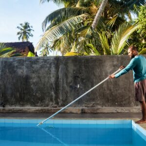 Cleaner man cleaning swimpool of fallen leaves with with vacuum equipment on backyard countryside