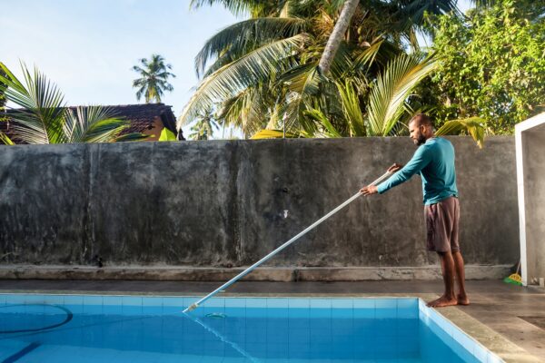 Cleaner man cleaning swimpool of fallen leaves with with vacuum equipment on backyard countryside